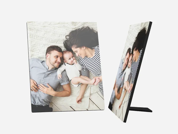 Ein Fotoaufsteller zeigt ein liebevolles Familienbild. Ein junger Mann, eine junge Frau mit lockigen, dunklen Haaren und ein kleines Baby liegen zusammen auf einer hellen, gestrickten Decke. Der Mann trägt ein hellblaues Hemd und schaut lächelnd zum Baby. 